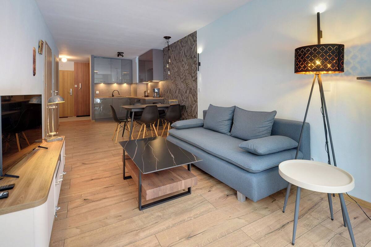 Una sala de estar moderna con un sofá gris, una mesa de centro y una lámpara de pie. Se observa una cocina al fondo con armarios de madera y un comedor con mesa y sillas.