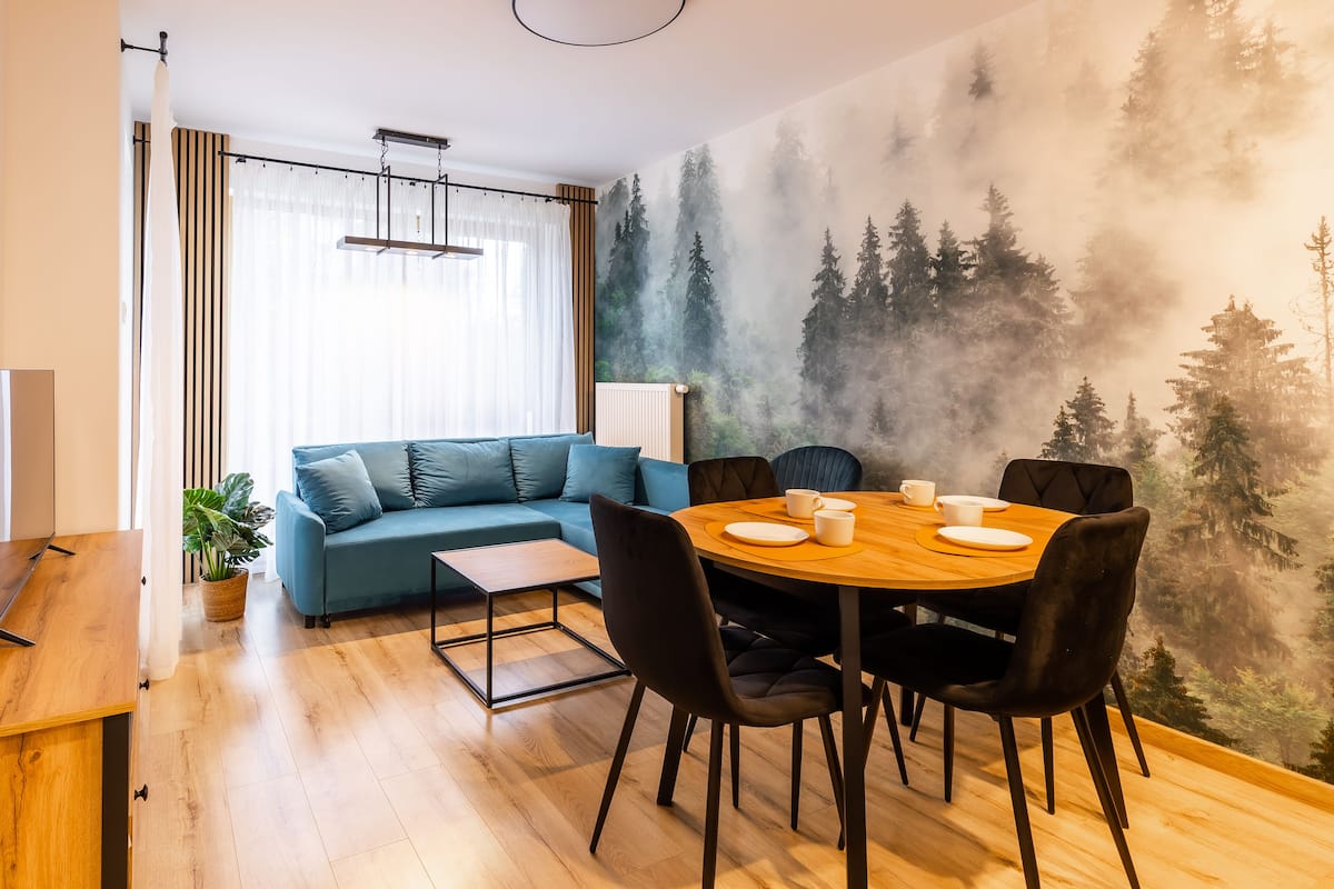 Una sala de estar moderna con un sofá azul, una mesa de comedor de madera con sillas negras y un mural con motivos de bosque en la pared.