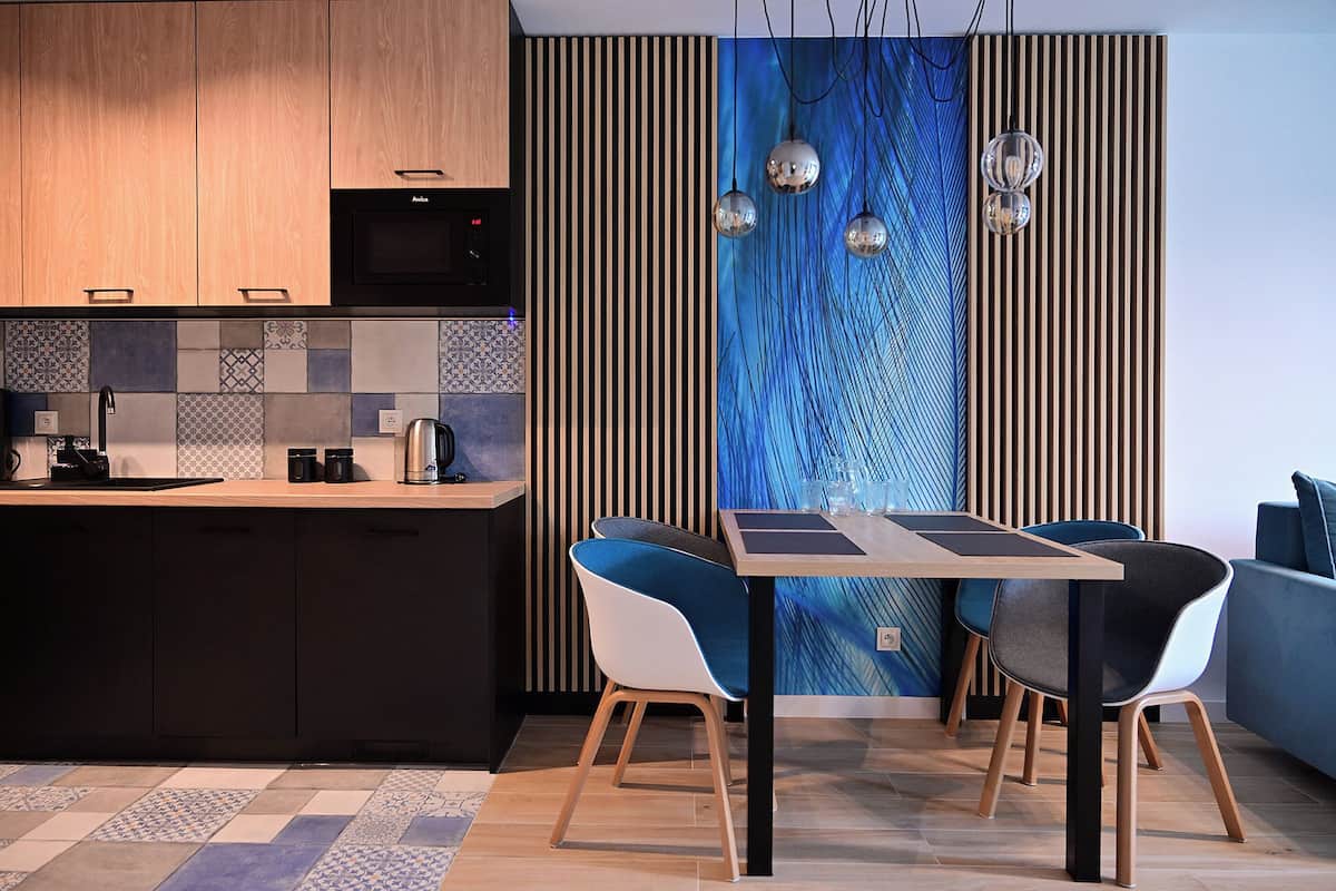 Una cocina moderna con encimera de madera, microondas integrado y hervidor. Hay una zona de comedor con mesa y sillas, y una pared decorativa con listones verticales de madera y arte abstracto azul.
