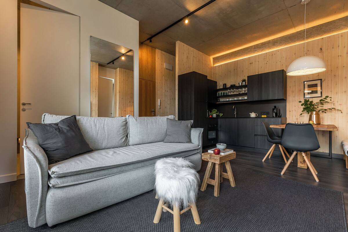 Una sala moderna con un sofá gris, un taburete pequeño y una mesa de comedor de madera con sillas.