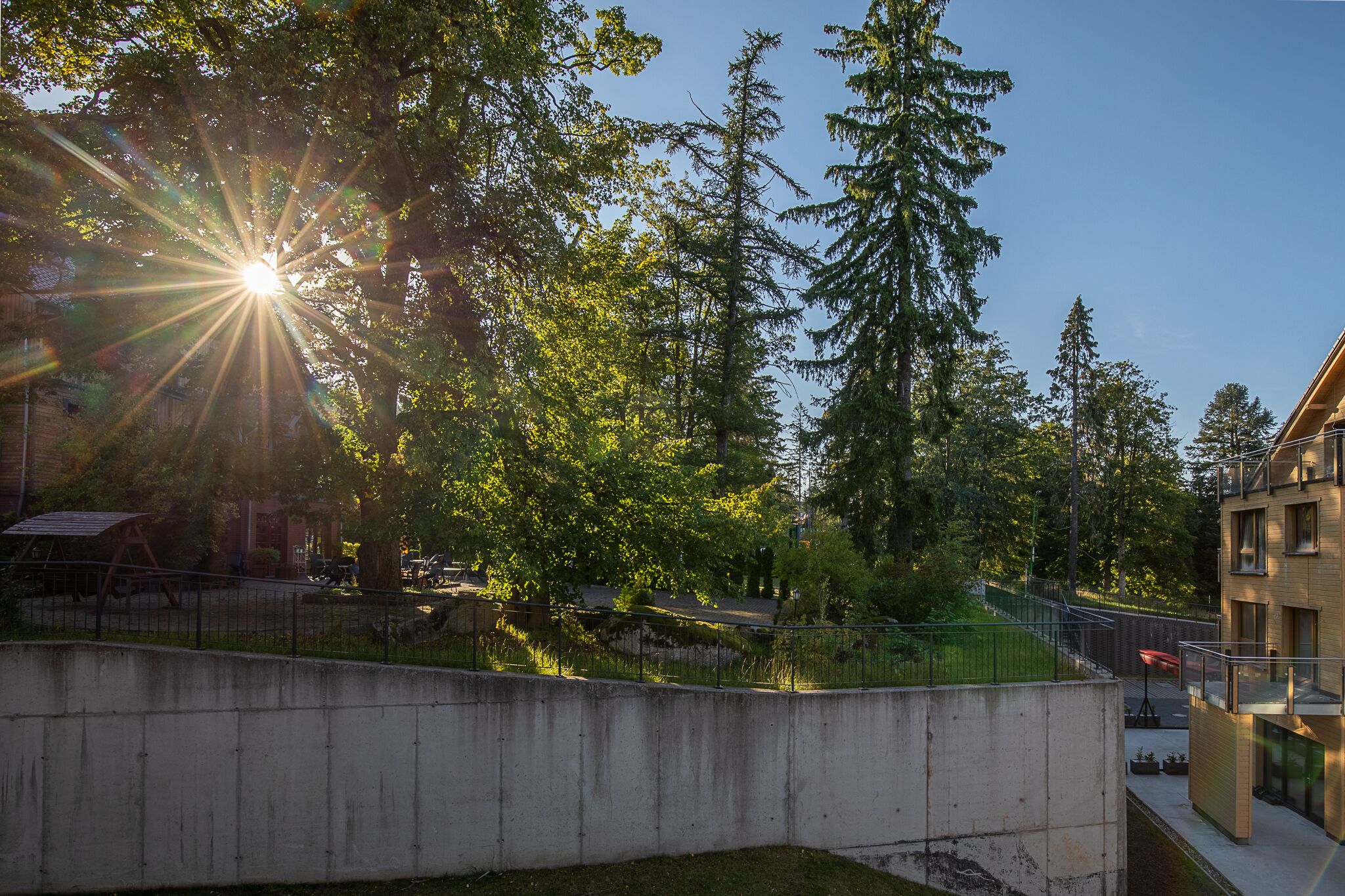 A concrete wall with a metal fence, trees, and a building with a balcony.