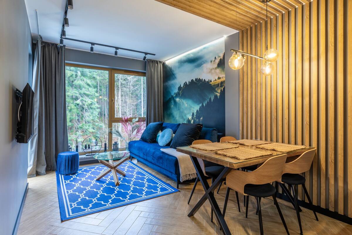 Un salón moderno con un sofá azul, una mesa de comedor de vidrio y un ventanal con vistas a árboles.