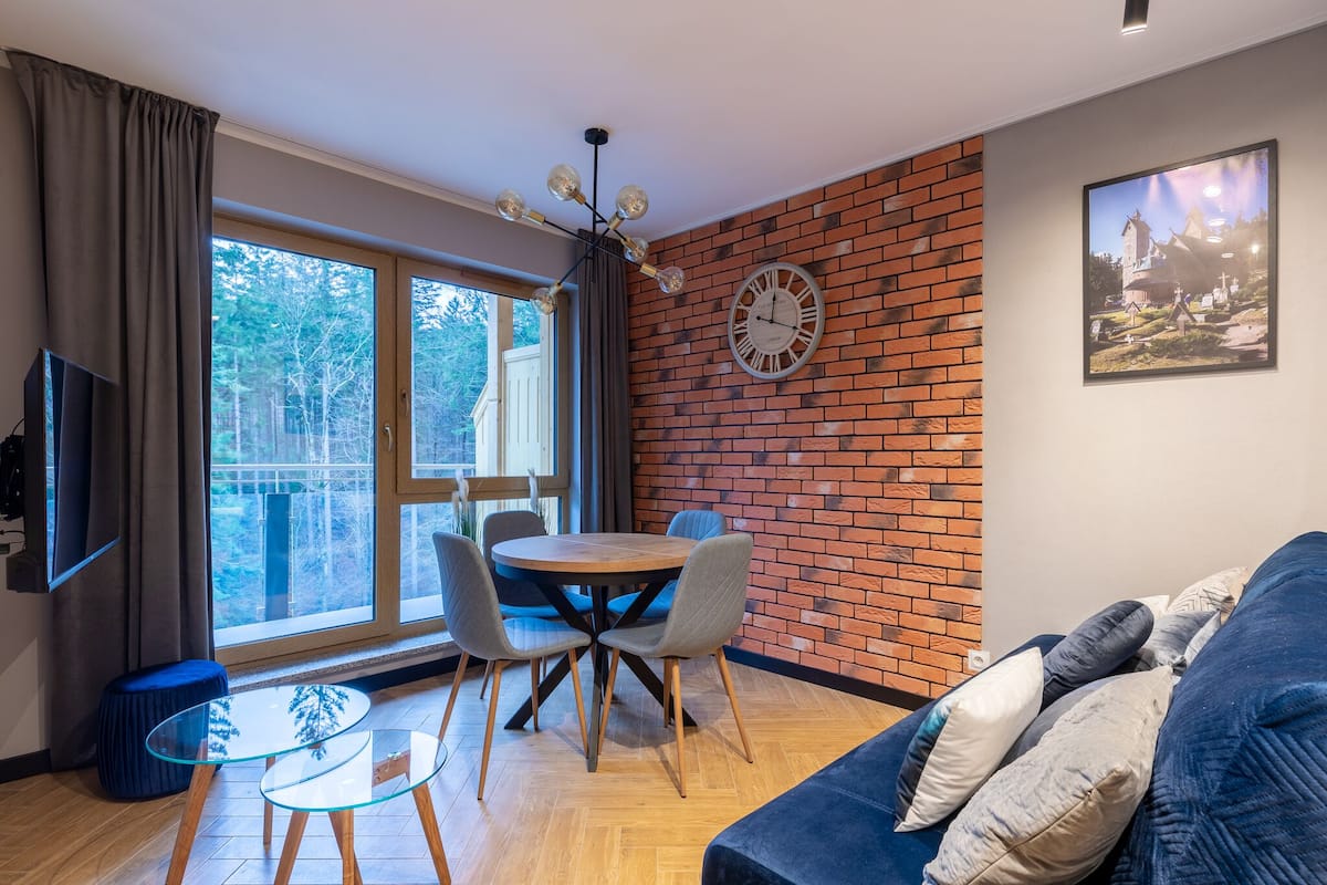 Una sala de estar moderna con una pared de ladrillo, una mesa de comedor redonda con sillas, un sofá azul y una televisión.