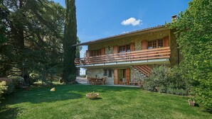 A two-story stone building with a wooden balcony, surrounded by lush greenery and a well-maintained lawn.