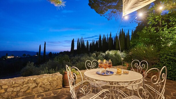 An outdoor dining area with a round table and chairs, surrounded by greenery and overlooking a landscape with trees and distant lights.