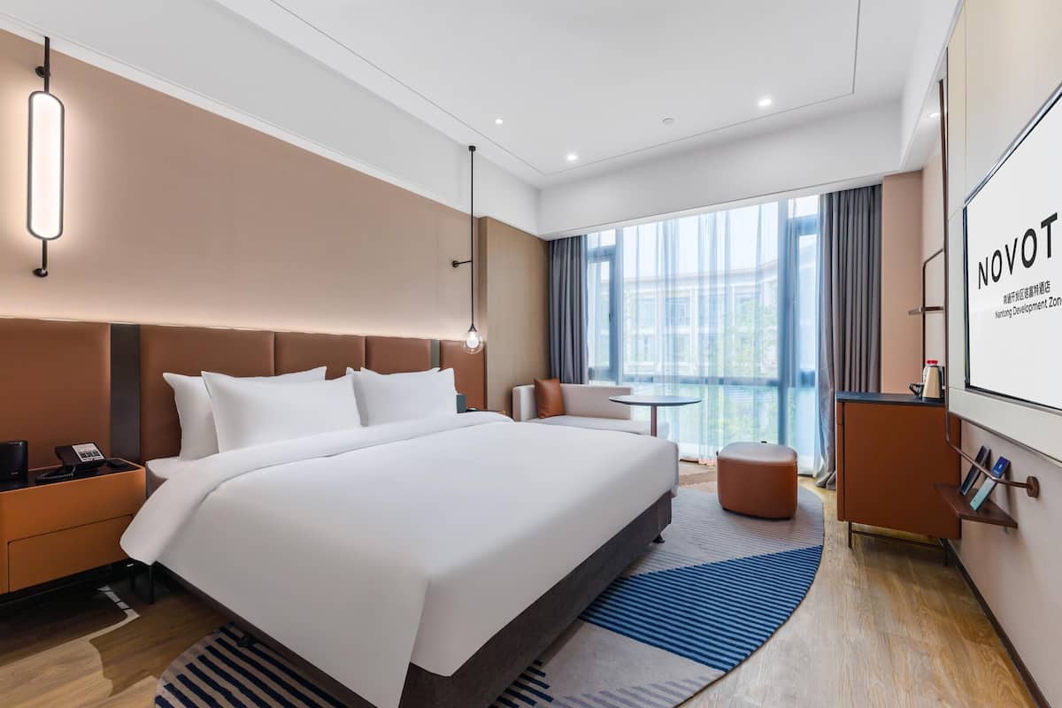 A modern hotel room with a large bed, a desk, a chair, a small table, and a balcony with a view.