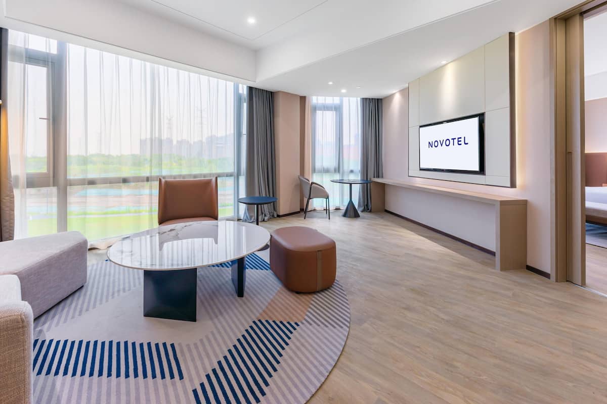 A modern hotel room with a large window, a round table, a sofa, and a TV screen displaying "NOVOTEL".