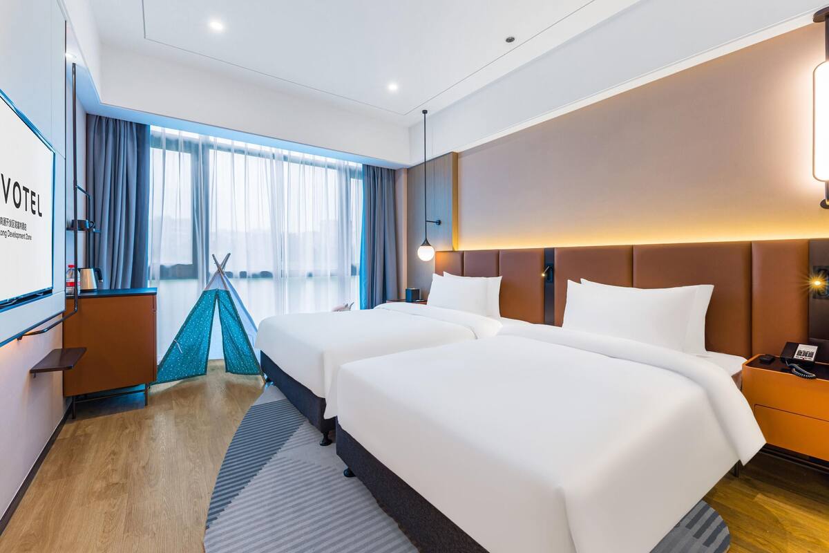 A hotel room with two beds, a desk, a chair, a large window with curtains, a wall-mounted lamp, and a teepee decoration.