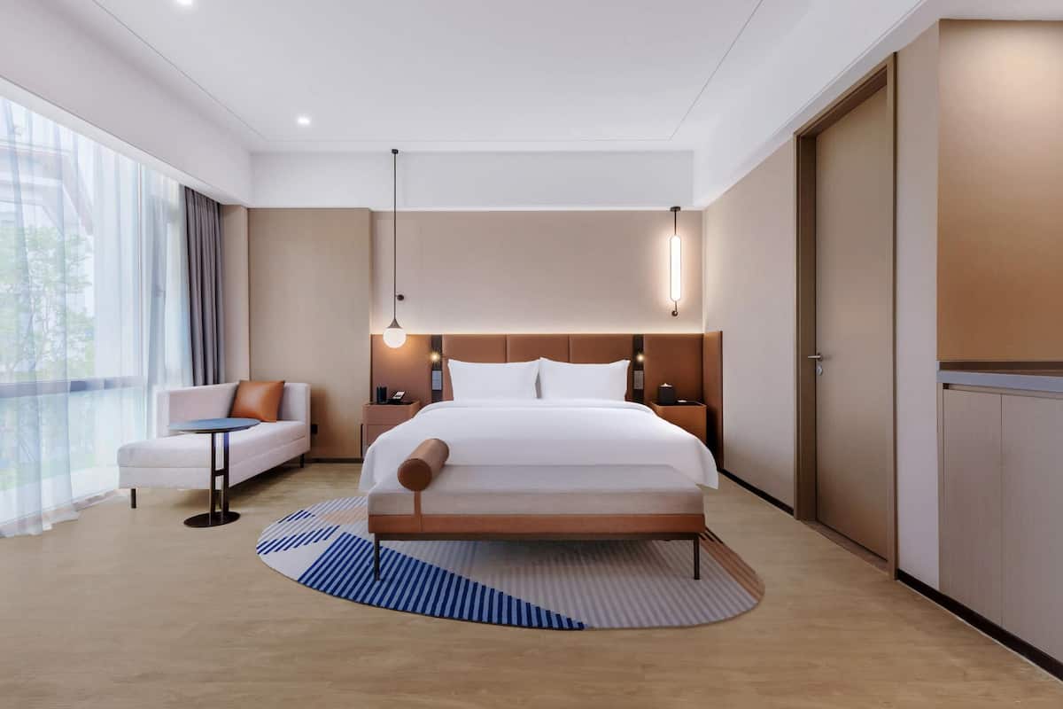 A modern hotel room with a large bed, a sofa, a small table, and a view of the outdoors.