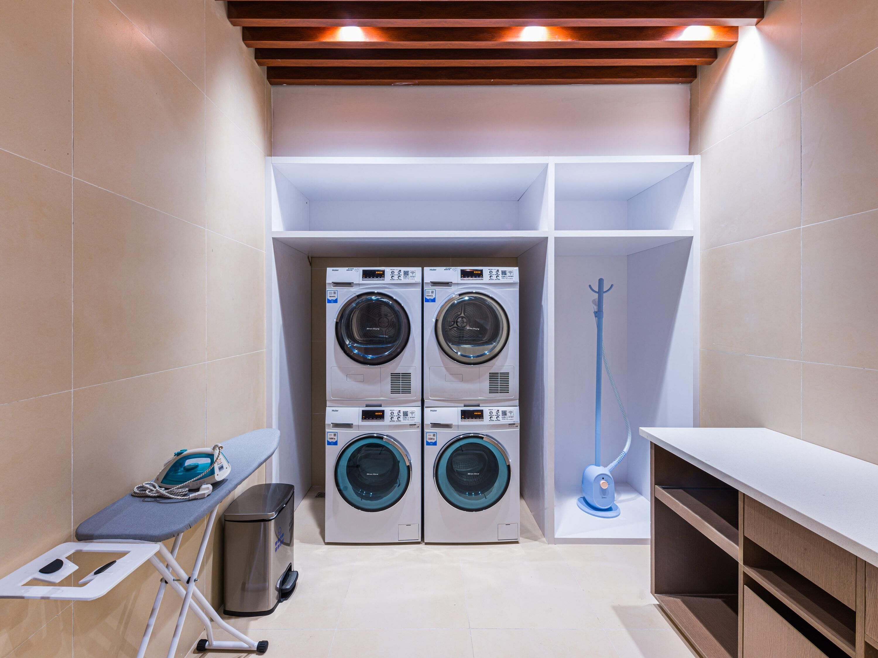 A laundry room with four stacked front-loading washing machines, a drying machine, an ironing station, and a blue handheld steamer.