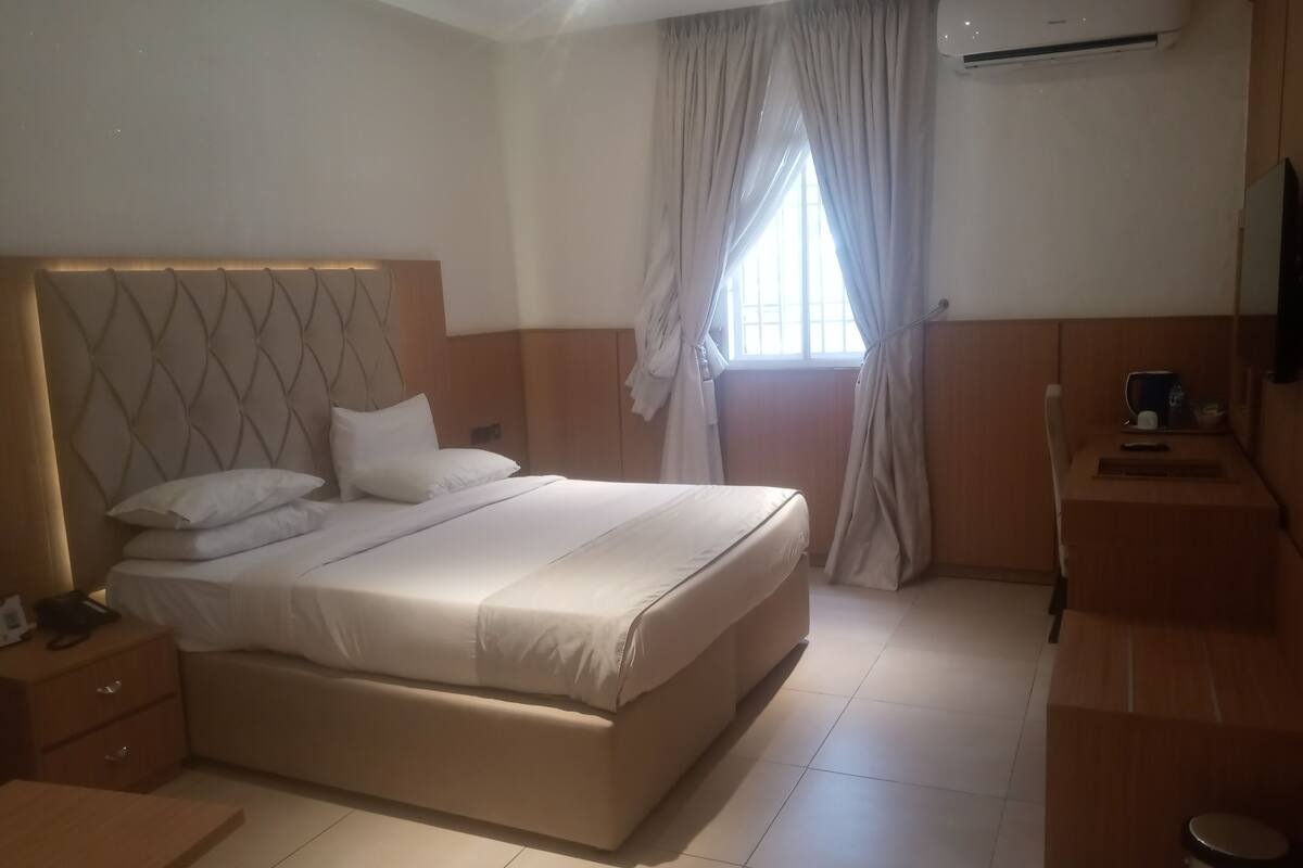 A hotel room with a large bed, a wooden headboard, a bedside table with a lamp, a small desk with a chair, and an air conditioning unit on the wall.