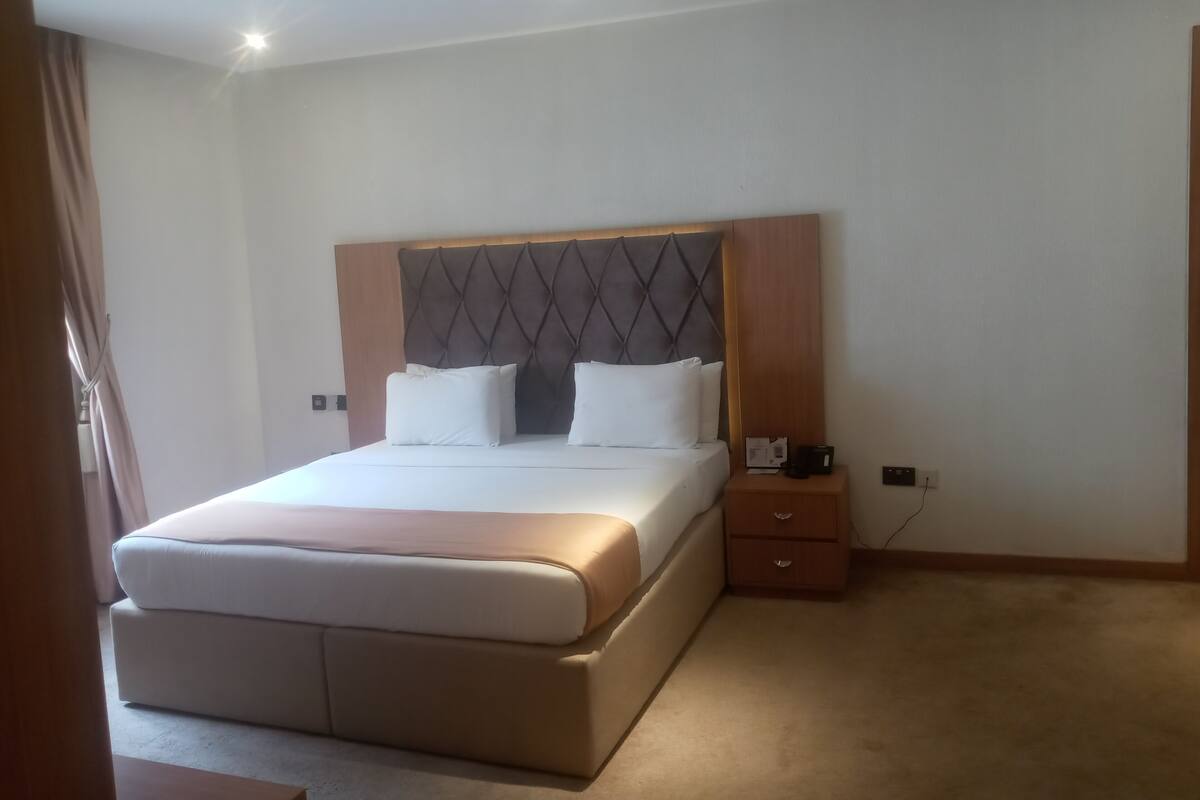 A hotel room with a large bed, a wooden headboard, a bedside table with a phone and a notepad, and a carpeted floor.