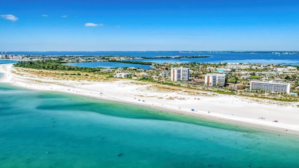 An aerial view of a coastal area with a beach, clear blue waters, and several high-rise buildings.