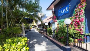A café with a blue sign, surrounded by lush greenery and a paved walkway.