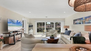 A living room with a TV, sofa, chairs, and a view of a residential area through large windows. - Kingston Arms - Sarasota (Sarasota)