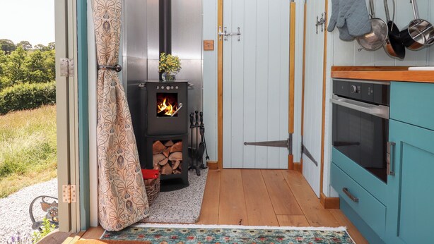 A cozy kitchen with a wood-burning stove, a dining area with a table and chairs, and a view of the outdoors. - Two Moors Shepherds Hut (Exeter)