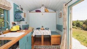 A compact kitchen with a sink, kettle, and toaster, a dining area with a table and chairs, and a view of greenery outside. - Two Moors Shepherds Hut (Exeter)