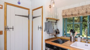 A kitchen with wooden cabinets, a built-in oven, a sink, and a window with a view of greenery. - Two Moors Shepherds Hut (Exeter)