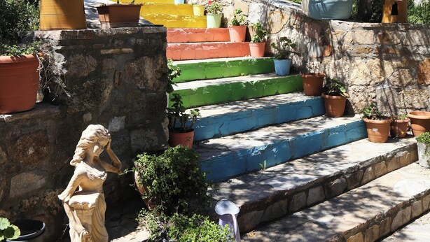 Una escala de pedra amb graons de colors que condueix a un edifici, envoltada de plantes en testos i una estàtua.