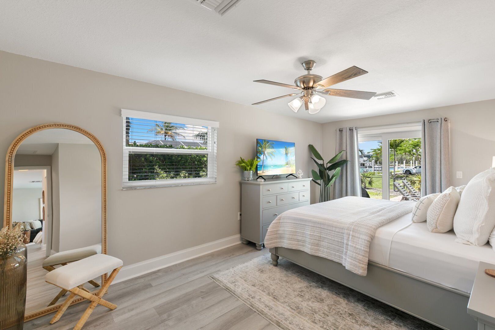 A bedroom with a bed, a ceiling fan, a large mirror, a dresser, and a window with a view of the outdoors.