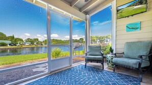 A room with large sliding glass doors, a blue patterned rug, two chairs, a small table with a plant, and a mounted TV displaying a golf course. - Lake View ~ Bayside Gated Resort ~ 2 Golf Carts (Miramar Beach)