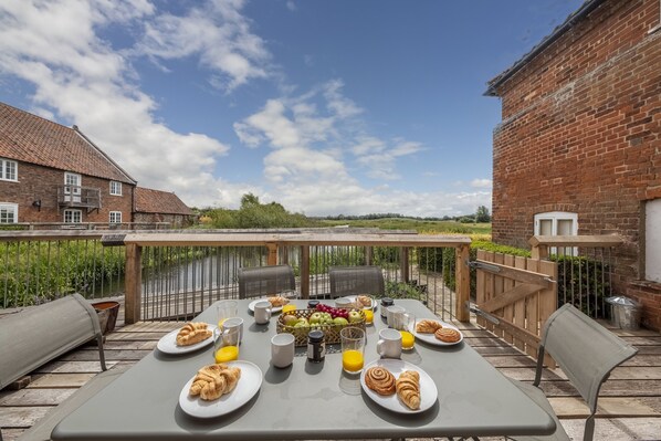 An outdoor dining area with a table set for breakfast, including croissants, fruit, and juice, overlooking a garden and a brick building.