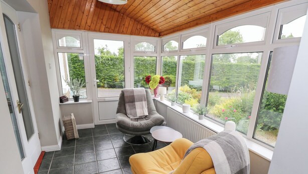 A sunroom with a wooden ceiling, large windows, a yellow chair, a grey chair, and a small table.