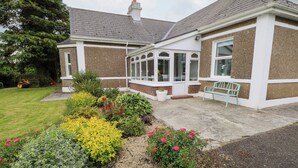 A house with a conservatory, a garden with various plants and flowers, and a bench outside.
