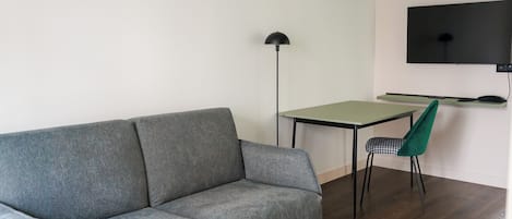 A living room with a grey sofa, a small table, a green chair, a floor lamp, and a TV mounted on the wall.