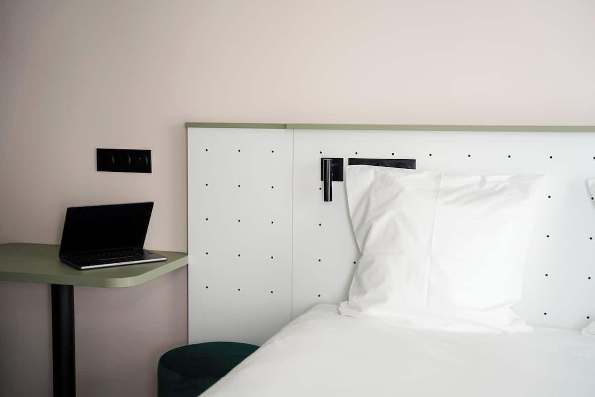 Hotellihuone, jossa on sänky, työpöytä kannettavalla tietokoneella ja seinälle kiinnitetty hylly.