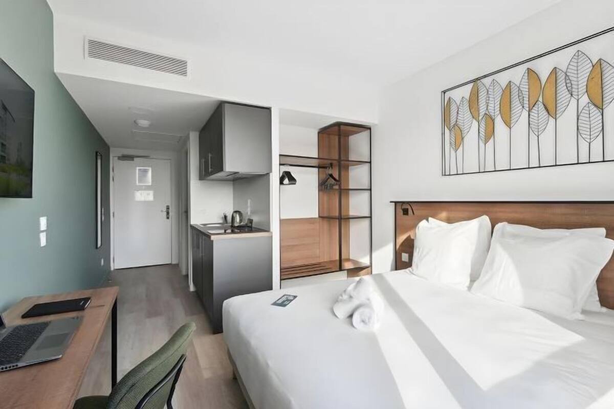 Moderni hotellihuone, jossa on sänky, työpöytä, pieni keittiö ja erillinen vaatehuone.