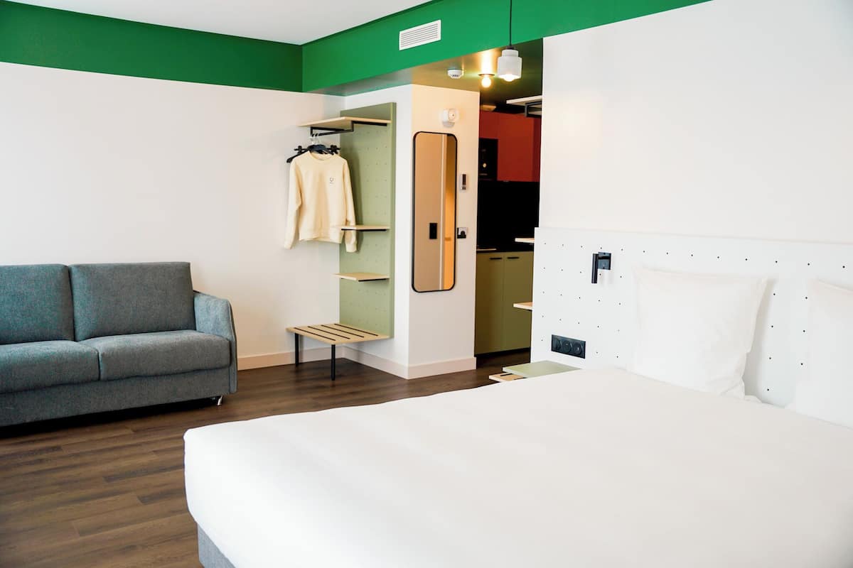 Moderni hotellihuone, jossa on harmaa sohva, suuri sänky, vaatekaappi ja pieni penkki.