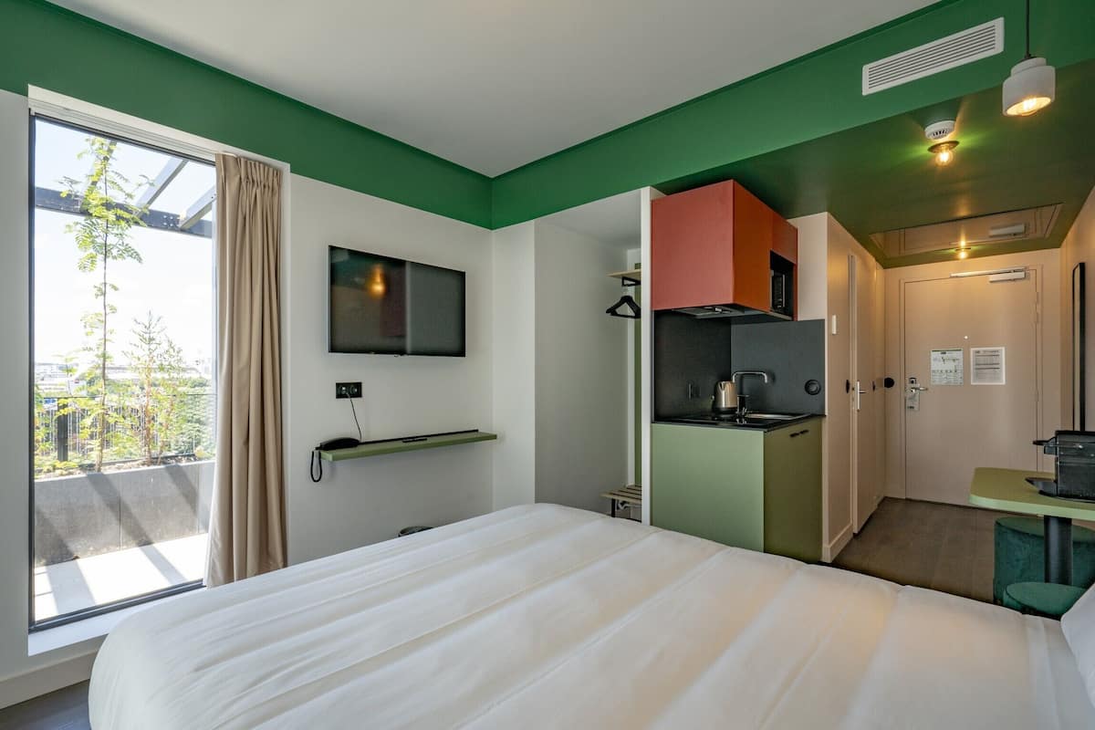 Hotellihuone, jossa on sänky, seinälle asennettu televisio, pieni keittiönurkkaus ja ikkuna, jossa on verhot.
