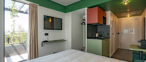 Quarto de hotel com cama, televisão presa na parede, cozinha compacta e janela com cortinas.