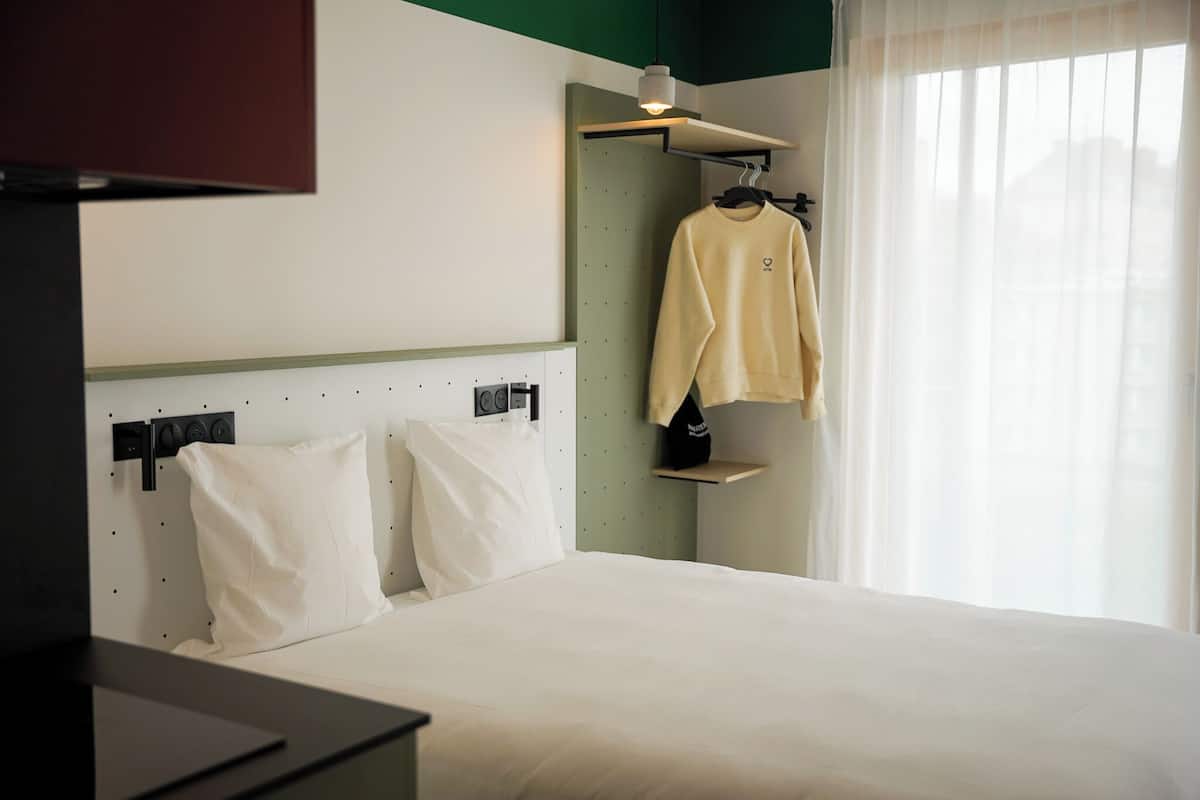 Hotellihuone, jossa on sänky, valkoiset tyynyt, riippuvalaisin, hylly, jossa on takki, ja ikkuna, jossa on läpikuultavat verhot.