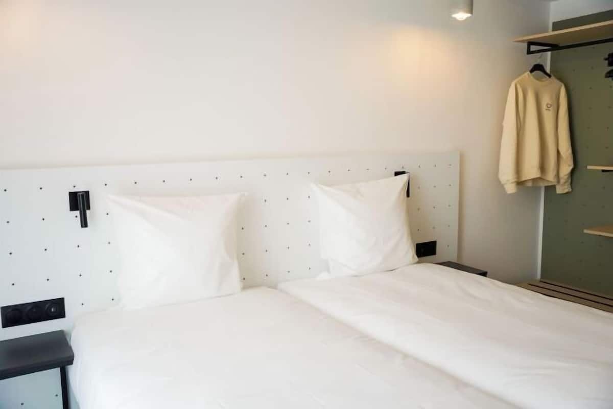 A bed with white bedding, a black nightstand, and a wall-mounted shelf with a hanging garment.