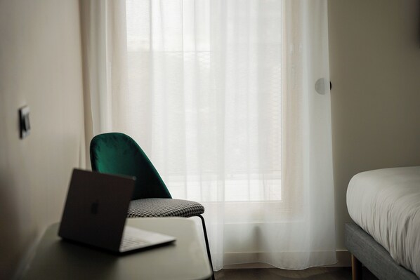 Una habitación de hotel con cama, escritorio, laptop, silla y ventana con cortinas ligeras.