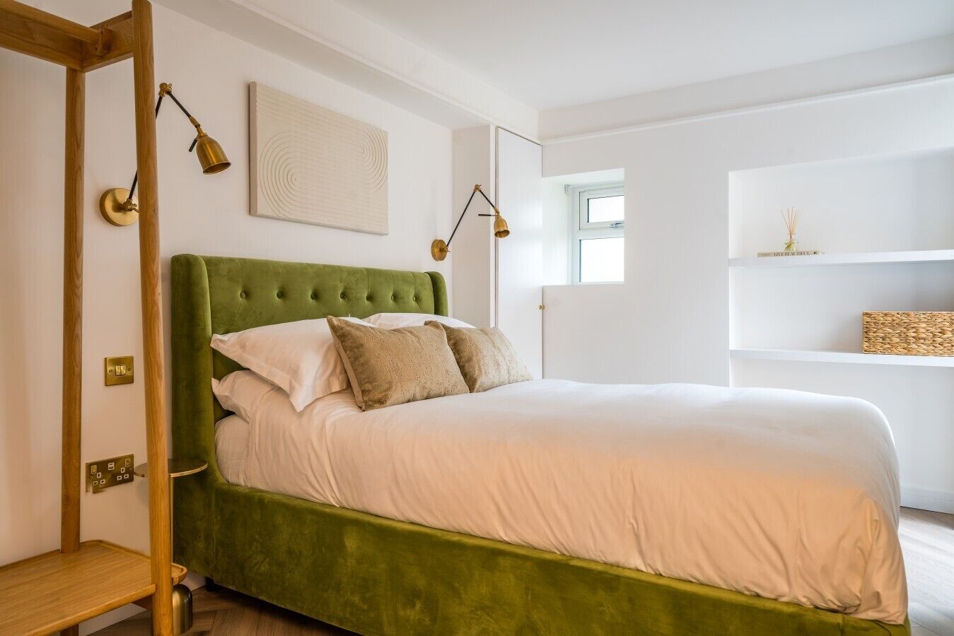 A bedroom with a green upholstered bed, white bedding, a bedside table, a wall-mounted lamp, and a shelf with decorative items.