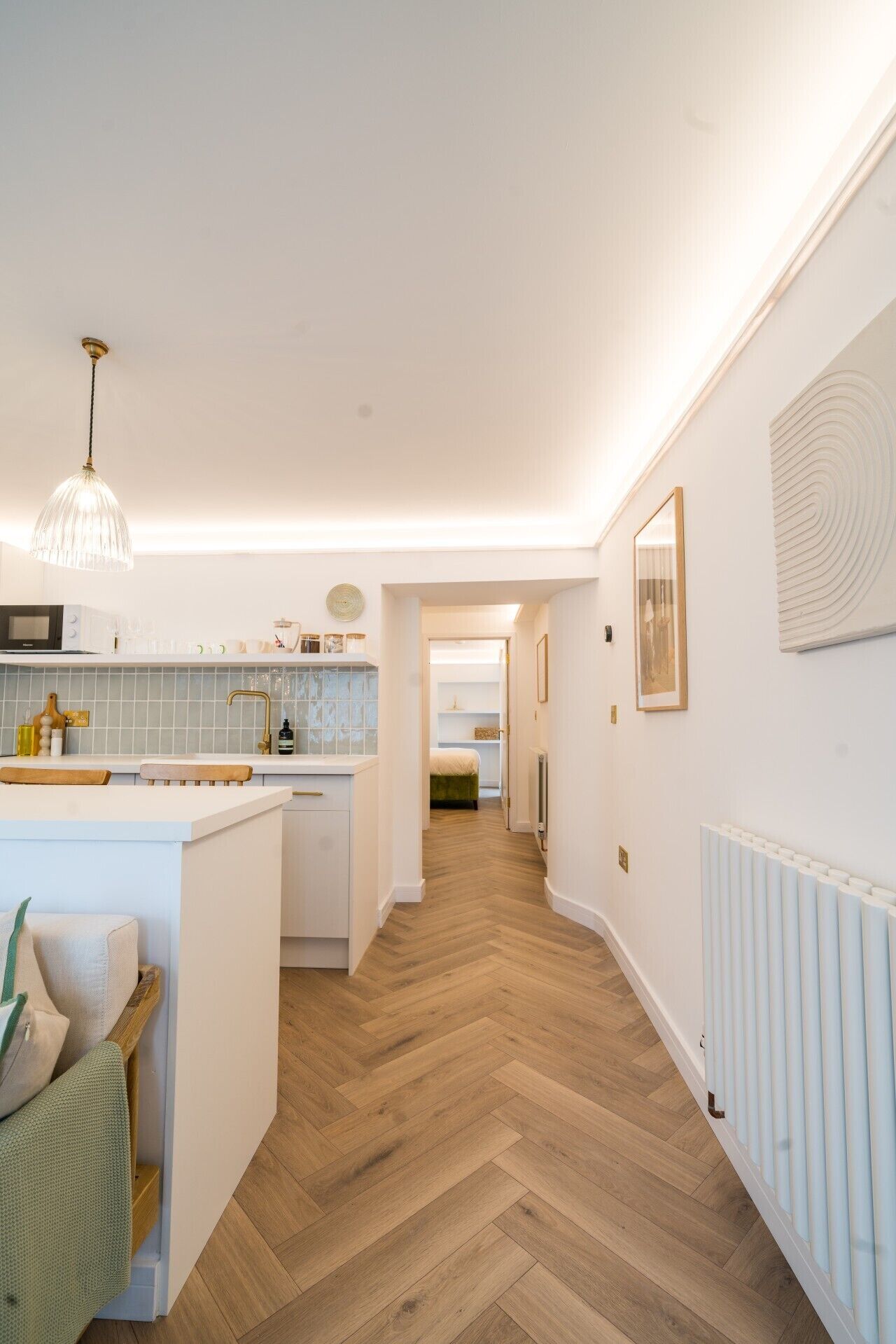 A modern kitchen with a central island, a dining area with a wooden bench, and a hallway leading to a bedroom.
