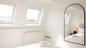 A bedroom with a skylight, a radiator, a bed with white linens, a bedside lamp, and a large mirror.