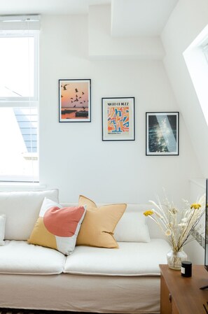 Ein modernes Wohnzimmer mit einem weißen Sofa, einem Couchtisch und einer Vase mit Trockenblumen. An der Wand hängen drei gerahmte Kunstwerke.