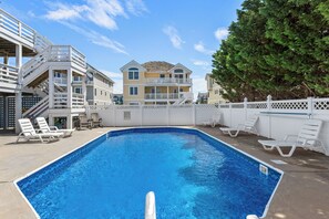 A swimming pool with white lounge chairs, a white fence, and a multi-story building in the background. - Captain Jack's Landing: Oceanside w/ Pool, Hot Tub (Nags Head)