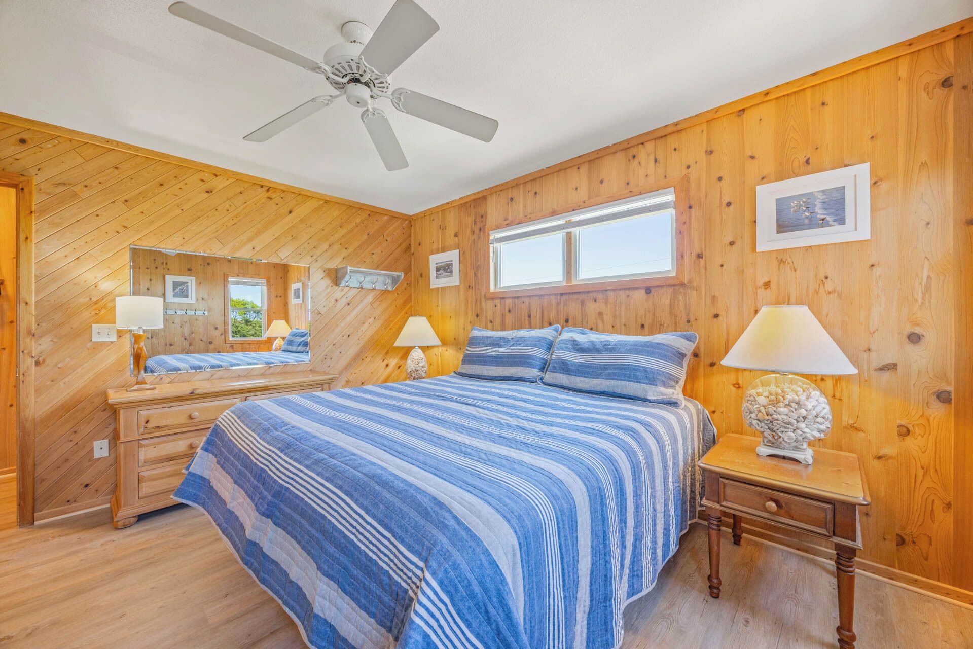 Unwind in your cozy bedroom featuring warm knotty pine walls, a comfortable bed with coastal-inspired linens, and natural light streaming through large windows. The ceiling fan keeps you cool on summer nights.