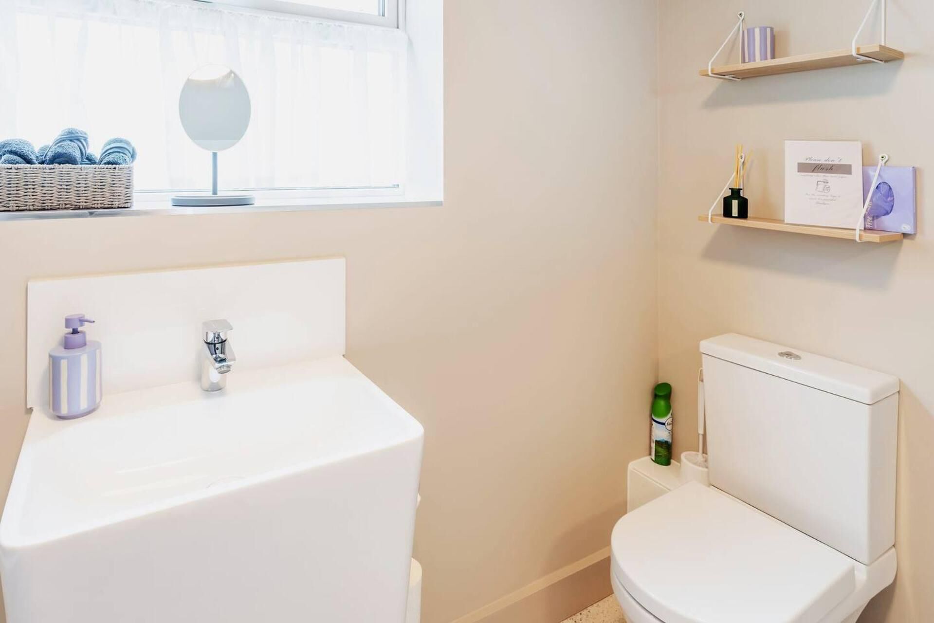 A bathroom with a white sink, a toilet, a window with a basket of towels, and a shelf with toiletries.