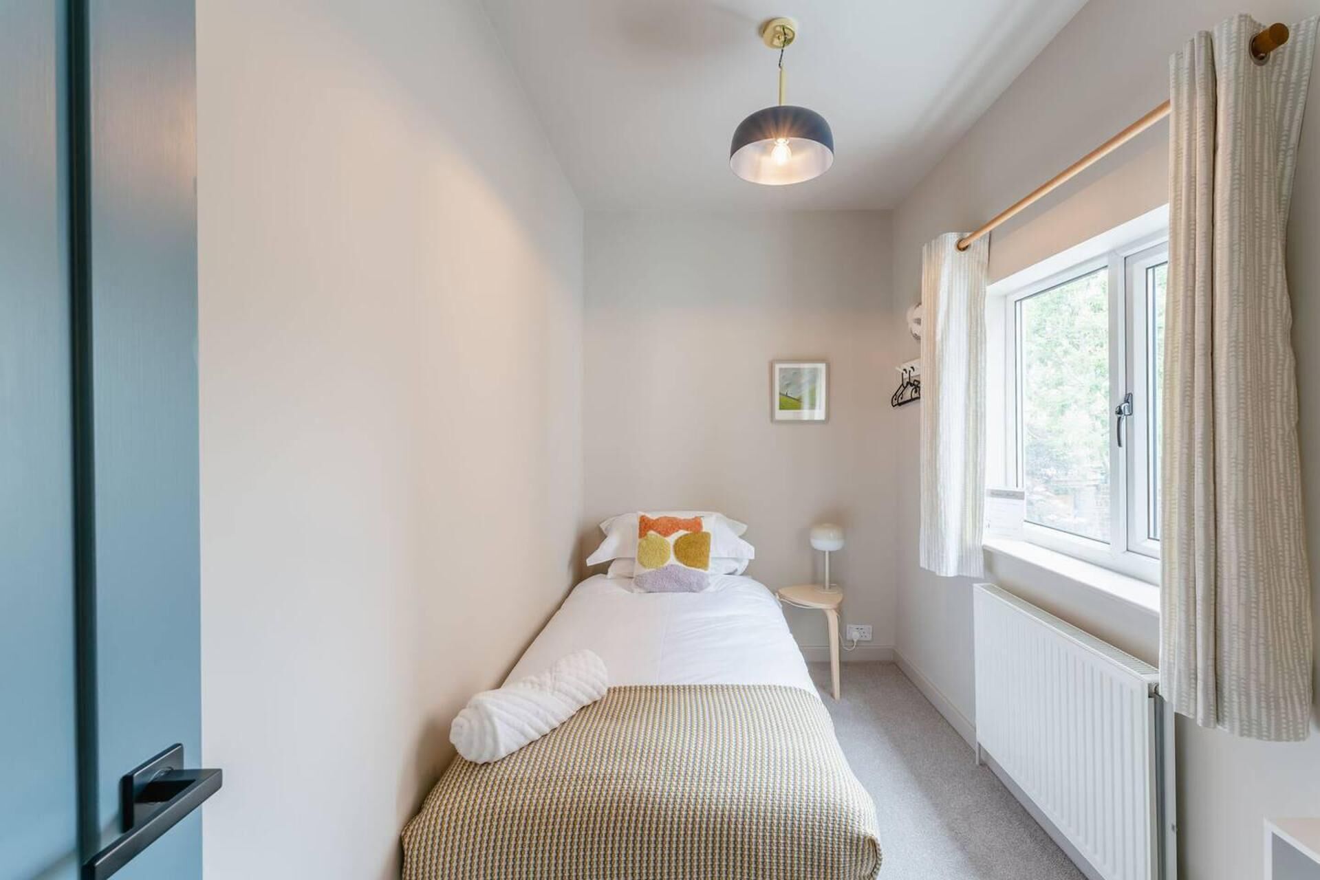 Et aflangt rum med en seng, et natbord, en hængende lampe og et vindue med gardiner.
