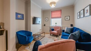 A living room with a blue sofa, a pink armchair, and a television on the wall. - Swan House 3-bed Town House York - up to 5 Guests (York)