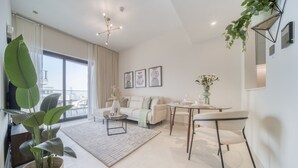 A modern living room with a sofa, coffee table, and dining table. There is a large window with curtains, a potted plant, and wall art.
