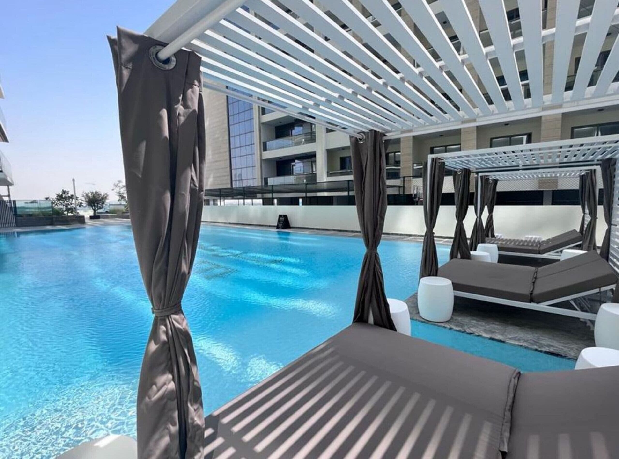 A pool area with a covered seating area, a clear blue pool, and a modern building in the background.