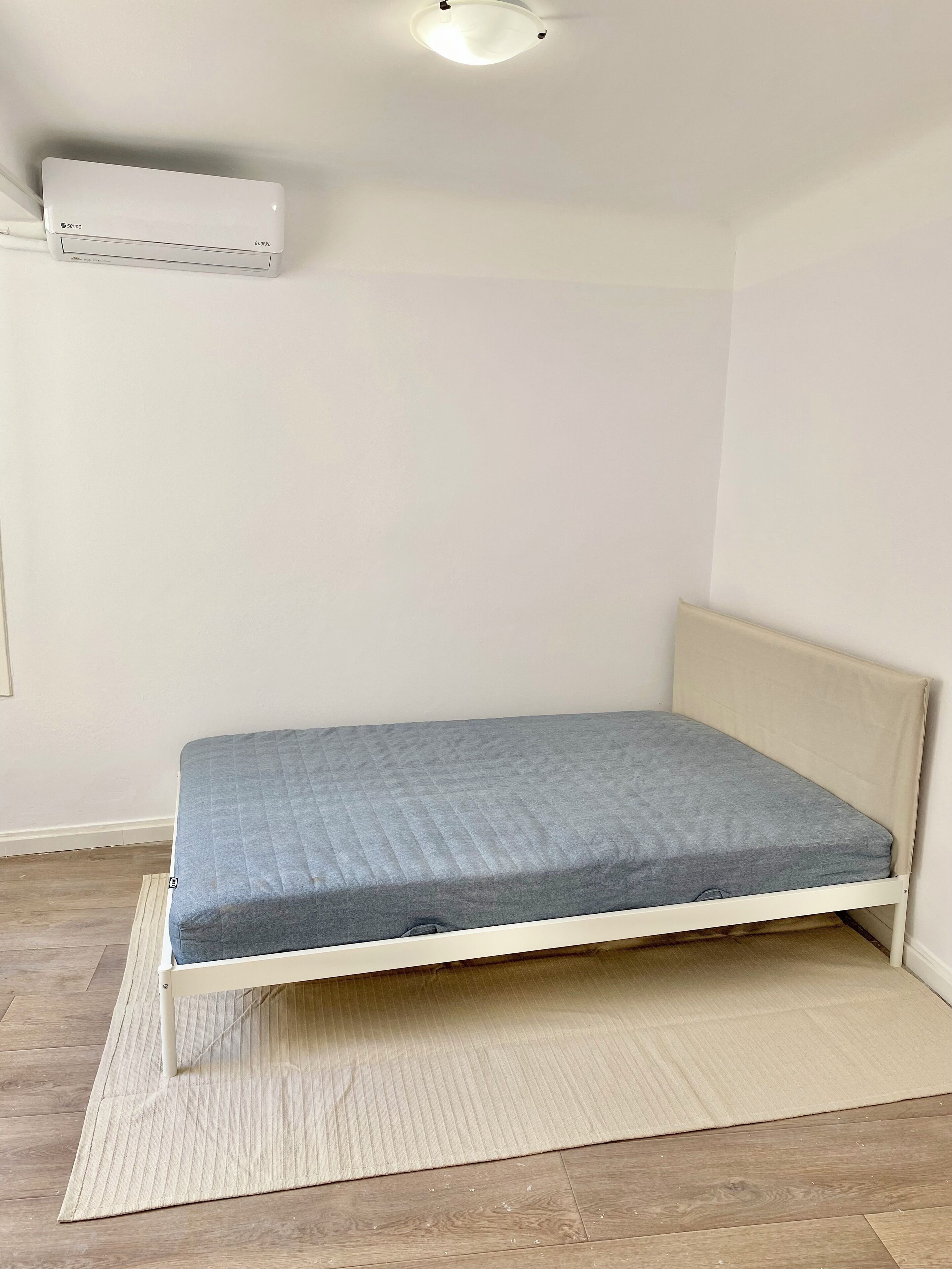 A small, single-bed room with a plain white wall, a ceiling-mounted air conditioner, and a wooden floor.
