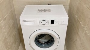 Una lavadora blanca de carga frontal, compacta, ubicada en una esquina de una habitación con paredes y piso de baldosas color beige.